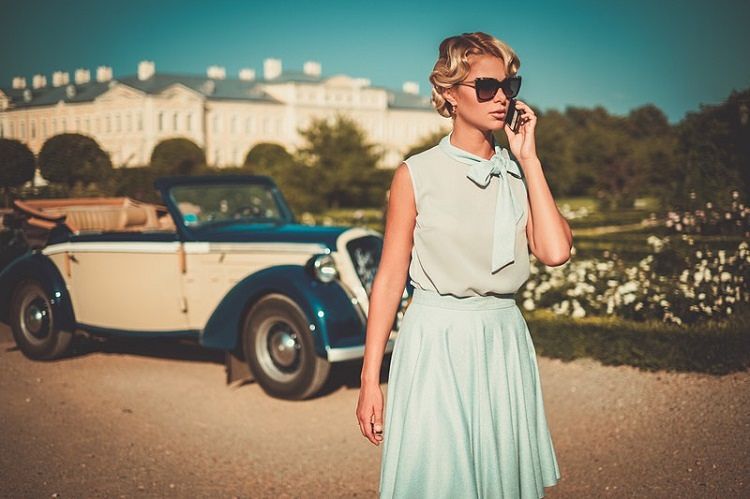 Mujer elegante con gafas de sol habla por el móvil junto a un coche clásico en un jardín